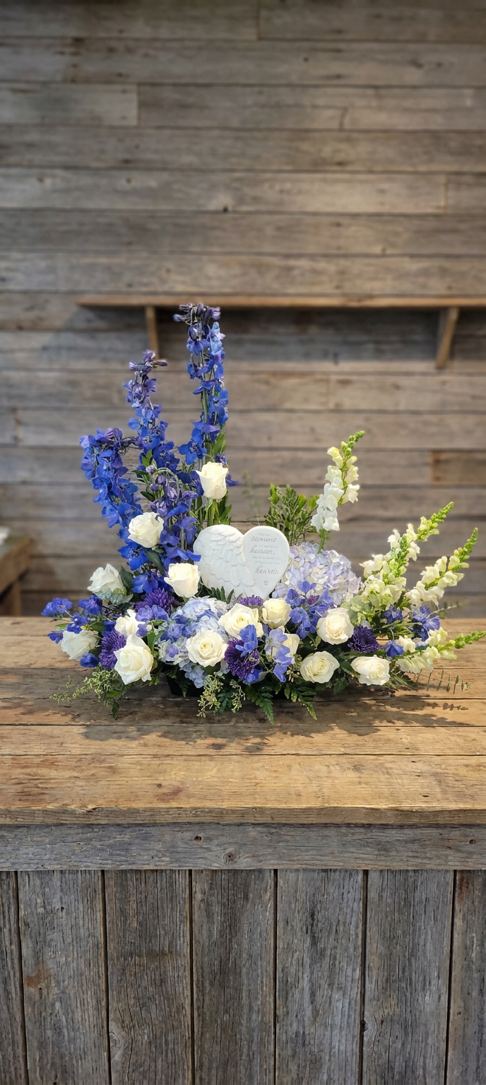 Asymmetrical floral arrangement of blue and white flowers with a central winged heart stone on a wooden table.