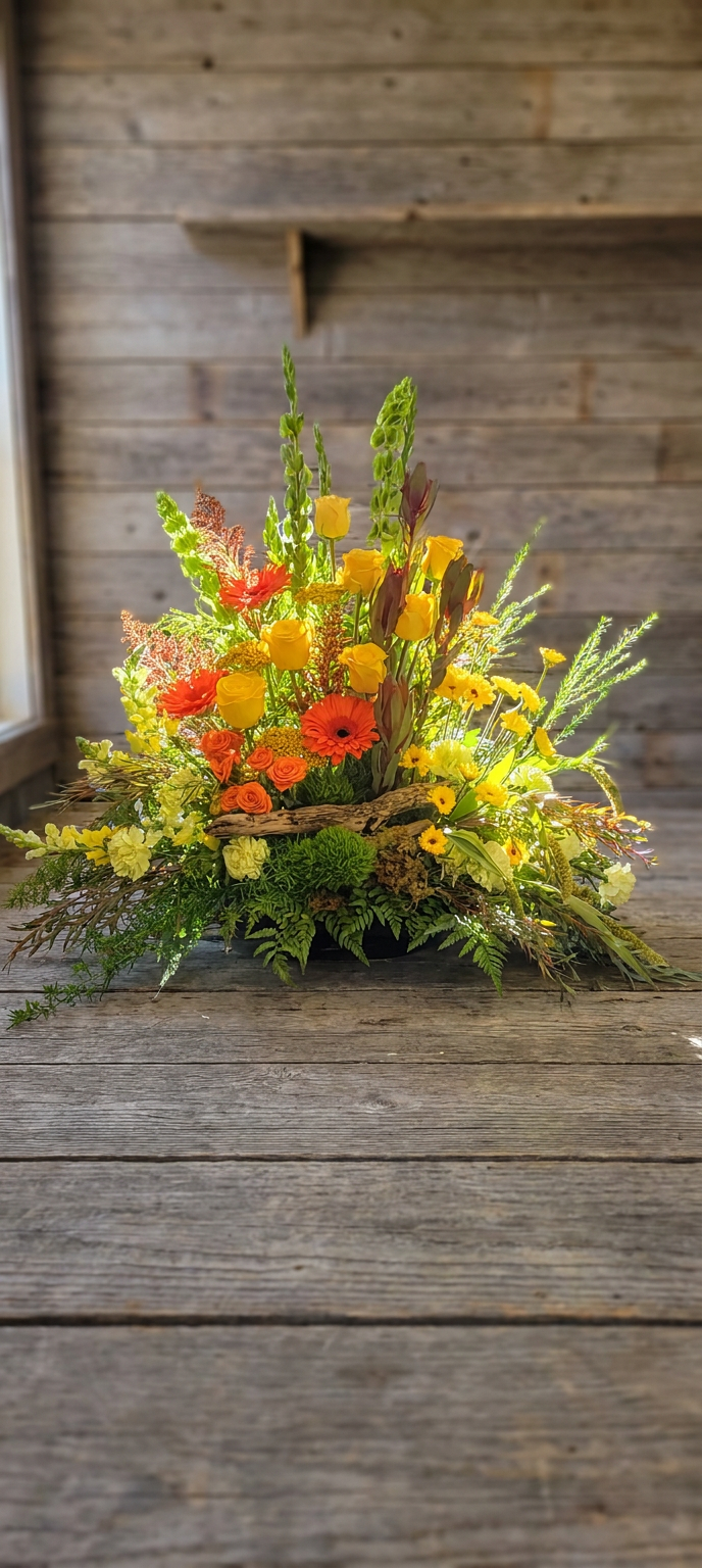 A large triangular arrangement of bright yellow and orange flowers with mixed greenery and natural wood.