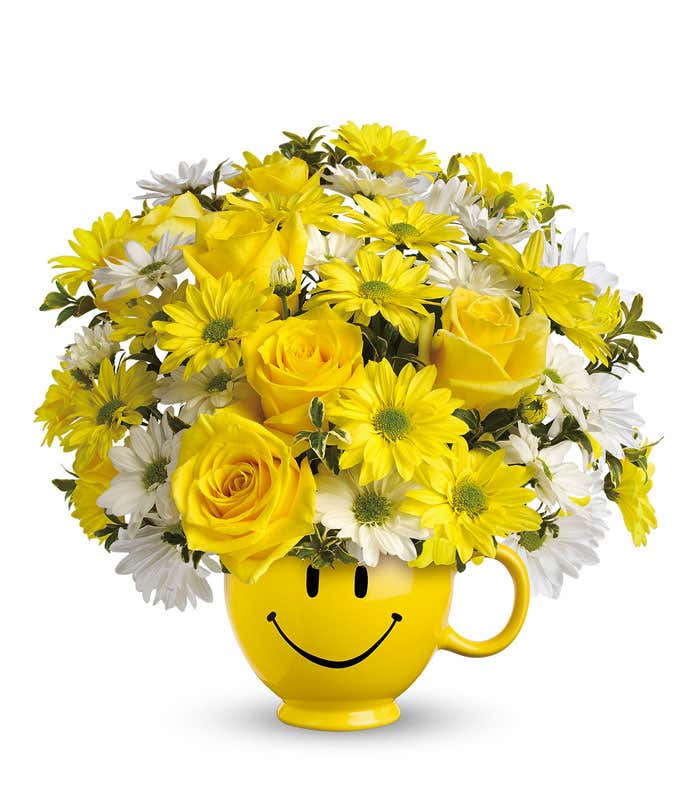 Yellow roses and daisies with white daisies arranged in a yellow ceramic smiley-face mug.
