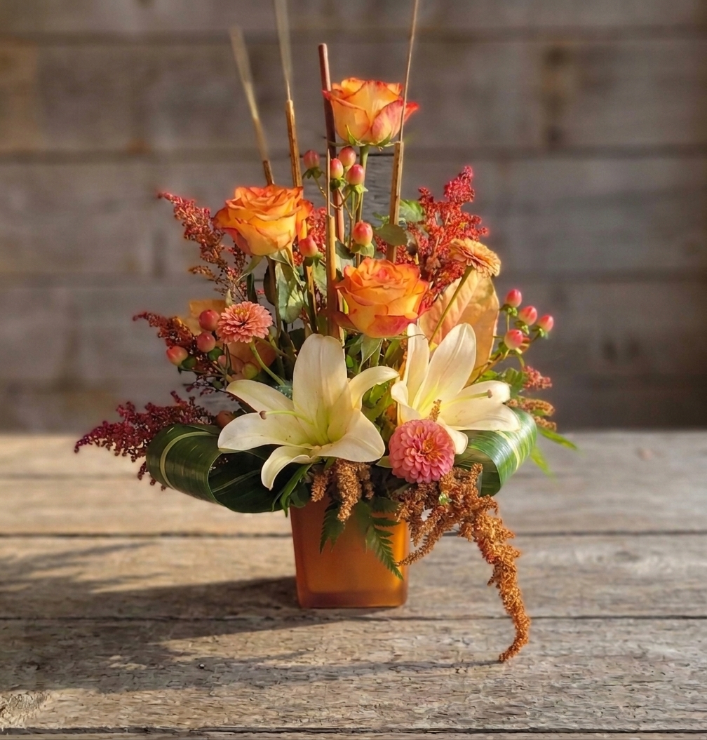 A full arrangement of warm orange roses and white lilies in a small amber glass cube.