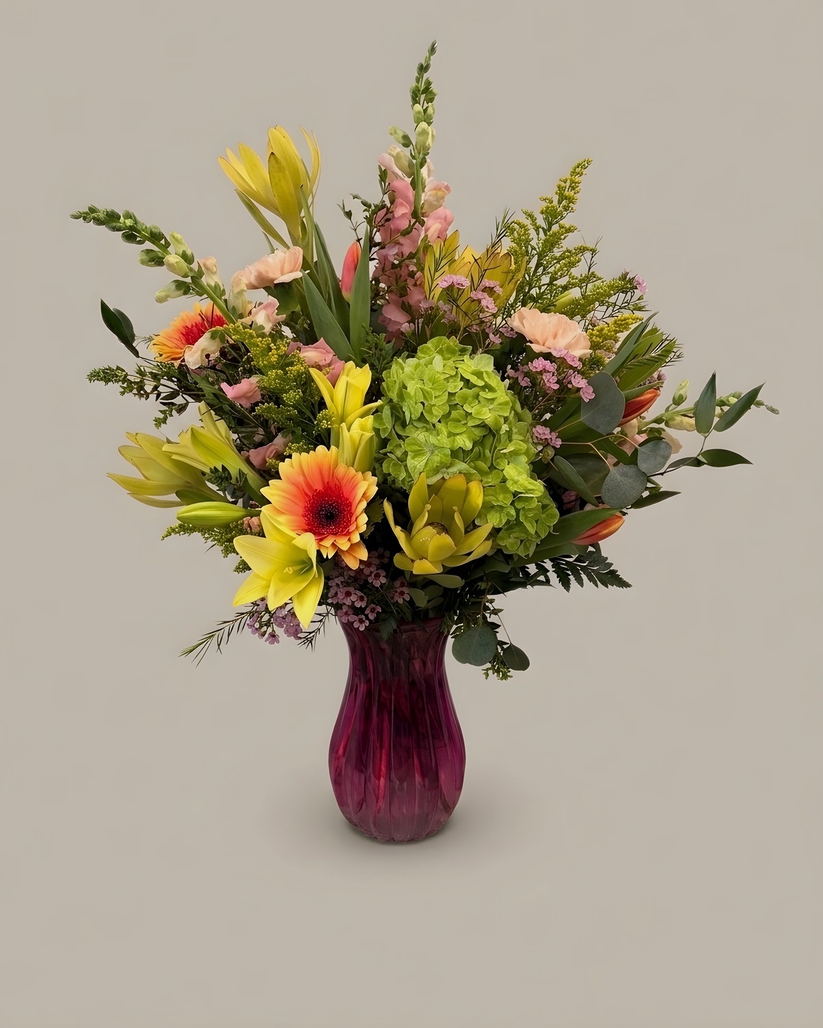 Green hydrangea and yellow snapdragon with a coral gerbera in a cranberry glass vase